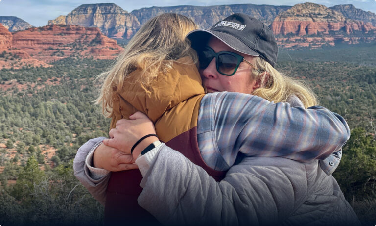 Two women hugging in front of a mountain range.